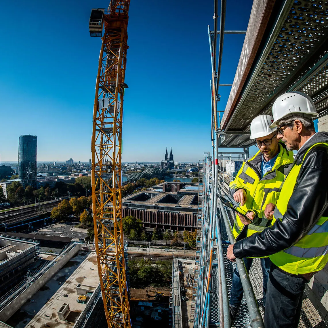 Zwei Bauleiter in Sicherheitskleidung besprechen Planungsunterlagen auf Gerüstplattform vor Kölner Skyline